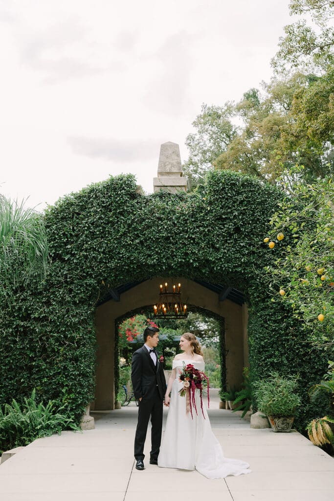 Sydonie Mansion Wedding Photos | Jennifer Holly Light and Airy Wedding Photographer in Orlando | Bride and groom stand in front of an ivy-covered archway. The bride holds a red bouquet. The atmosphere is romantic and serene, with lush greenery surrounding them.