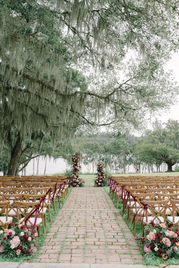 Sydonie Mansion Wedding Photos | Jennifer Holly Light and Airy Wedding Photographer in Orlando | Outdoor wedding venue with brick aisle lined by wooden chairs and floral arrangements. Large oak tree with hanging moss creates a serene backdrop.