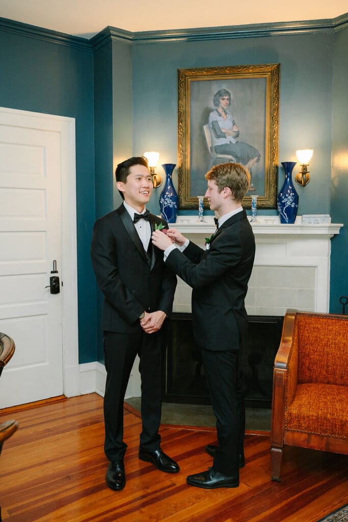 Sydonie Mansion Wedding Photos | Jennifer Holly Light and Airy Wedding Photographer in Orlando | Two men in formal suits stand in a warmly lit room with blue walls and wood floors. One adjusts the other's boutonniere, smiling. A classic portrait hangs above a mantel.
