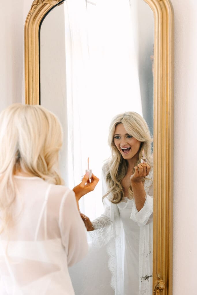 bride putting on lipgloss while looking in a gold mirror at a wedding at The Howey Mansion captured by Jennifer Holly Photography