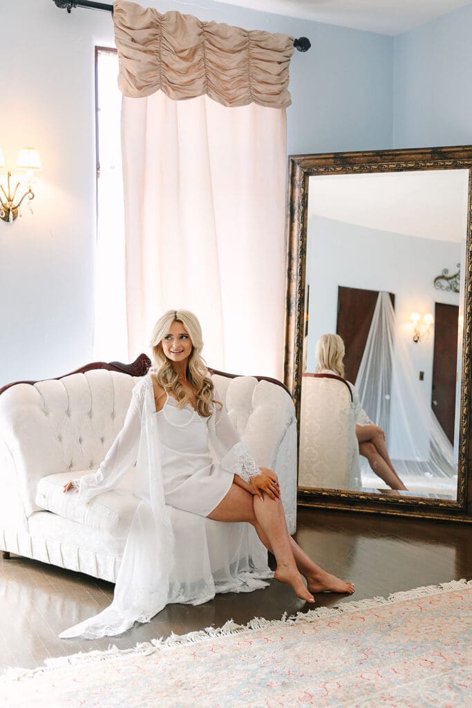 A bride in a white robe sits on a tufted sofa in a bright room at Howey Mansion, with a large ornate mirror reflecting her and sheer curtains framing this elegant Florida estate wedding venue.