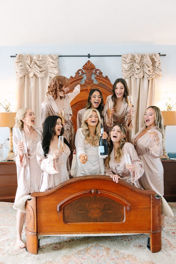 bride and bridesmaids in robes sitting and laughing on a bed with champagne at a wedding at The Howey Mansion captured by Jennifer Holly Photography