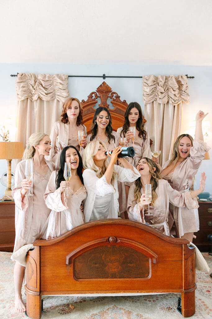 bride and bridesmaids in robes sitting and laughing on a bed with champagne at a wedding at The Howey Mansion captured by Jennifer Holly Photography