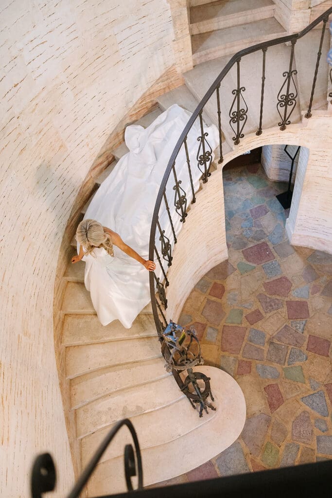 Bride in a white gown descends a curved stone staircase with a wrought iron railing at the historic Howey Mansion, a renowned Florida estate wedding venue, captured from above by Jennifer Holly Photography. Multicolored stone tiles adorn the floor below.