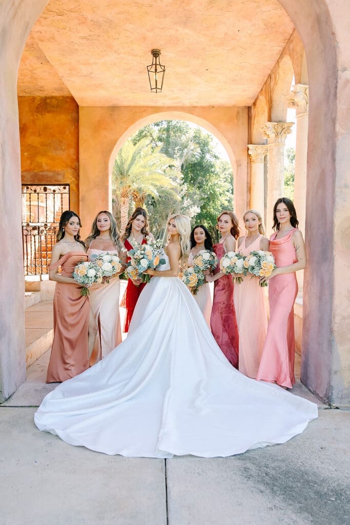 bride in white with bridesmaids in colors of the sunset under stone archways at The Howey Mansion captured by Jennifer Holly Photography