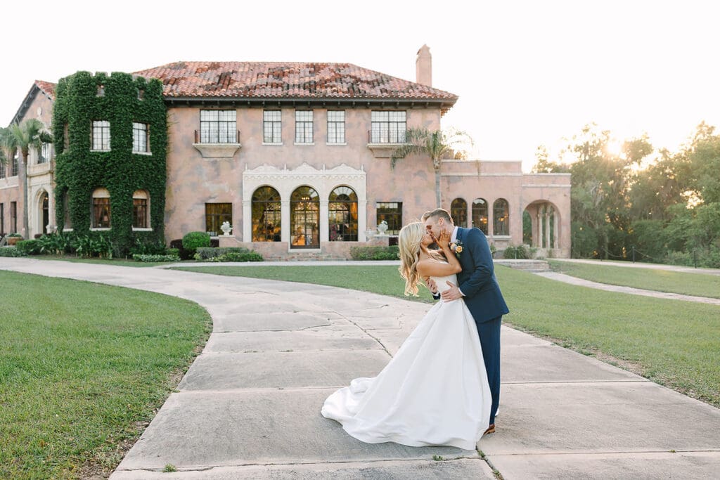 A bride and groom kiss while posing on a curved walkway at a Howey Mansion wedding during sunset, captured by Jennifer Holly Photography at this stunning Florida estate wedding venue.