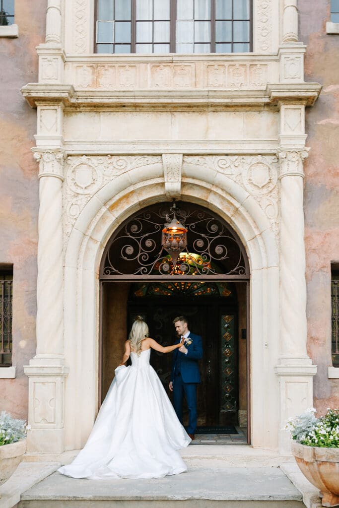 A bride in a white gown and a groom in a blue suit stand under an ornate stone archway at Howey Mansion, holding hands in front of a large wooden door at this stunning Florida estate wedding venue. Captured by Jennifer Holly Photography.