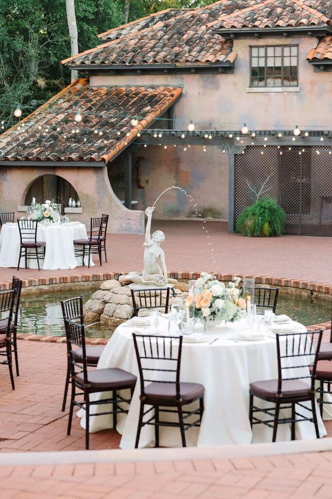 Outdoor courtyard with round tables set for an event, a small fountain and statue, string lights above, and the rustic Howey Mansion in the background—an idyllic Florida estate wedding venue. Captured by Jennifer Holly Photography.