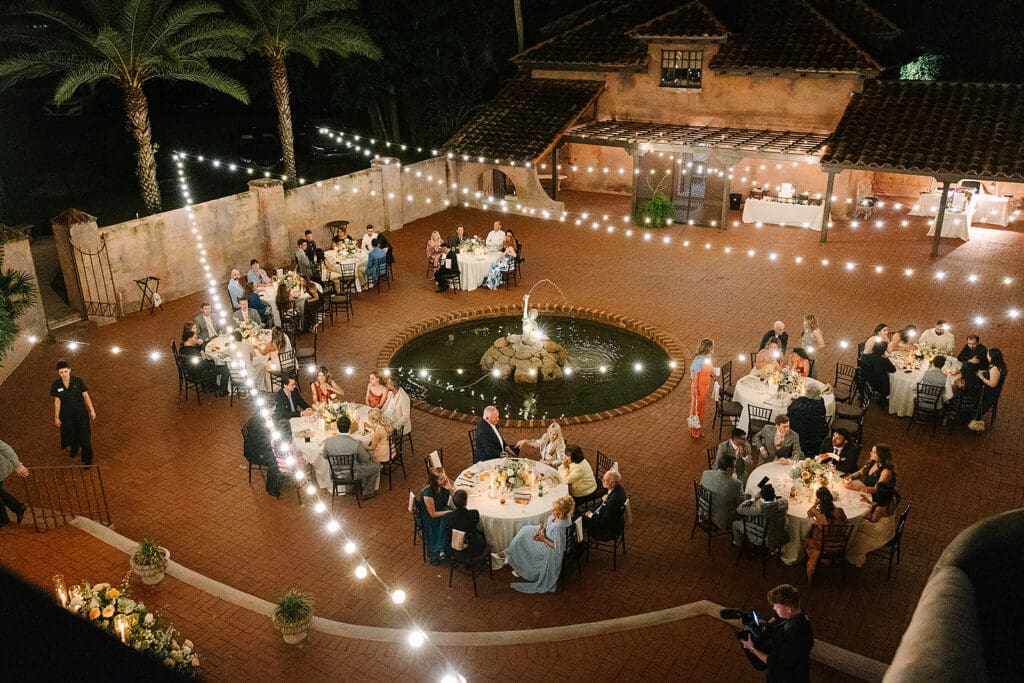 An outdoor evening wedding reception at the Howey Mansion, a stunning Florida estate wedding venue, with guests seated at round tables around a central fountain, string lights overhead, and a rustic building in the background. Captured by Jennifer Holly Photography.