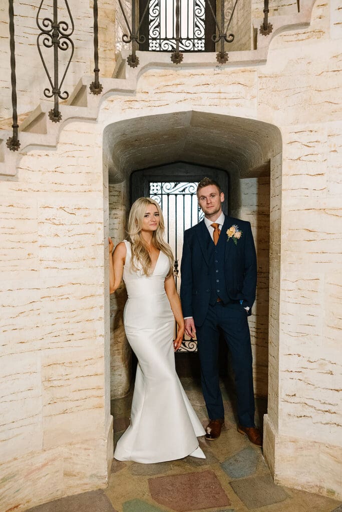 A bride in a white dress and a groom in a navy suit stand holding hands under a stone archway at the historic Howey Mansion, captured by Jennifer Holly Photography.